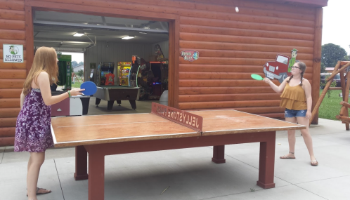 Two girls play table tennis outside a wooden building, with arcade games visible in the background.