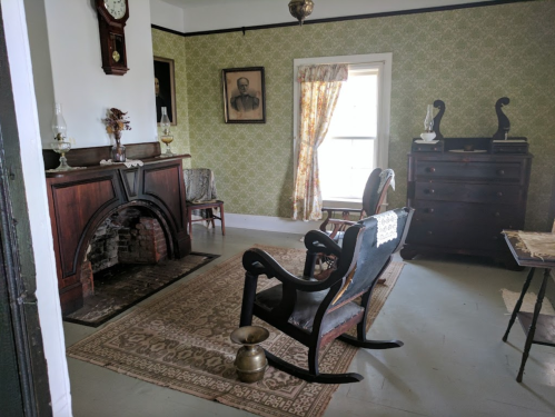 A vintage room with a rocking chair, fireplace, patterned wallpaper, and antique furniture, featuring a window with curtains.