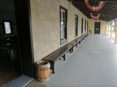 A porch with wooden benches, a trash can, and patriotic decorations, featuring a view of a grassy area outside.