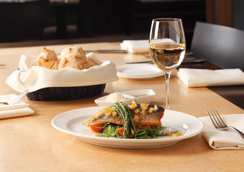 A plated fish dish with greens and tomatoes, accompanied by a glass of white wine and a basket of bread in the background.