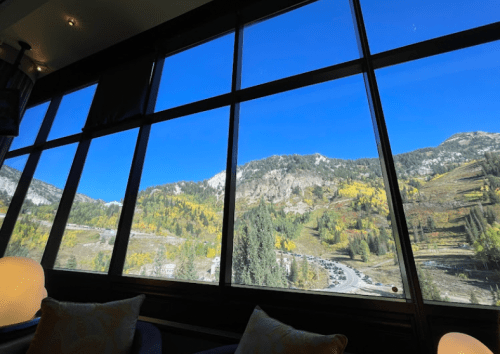 A scenic view of mountains and trees with autumn colors, seen through large windows against a clear blue sky.