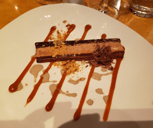 A rectangular chocolate dessert topped with mousse, chocolate shavings, and drizzled with caramel sauce on a white plate.