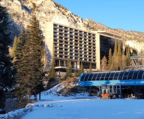 A modern hotel building surrounded by snow-covered trees and mountains, with a ski lift station in the foreground.