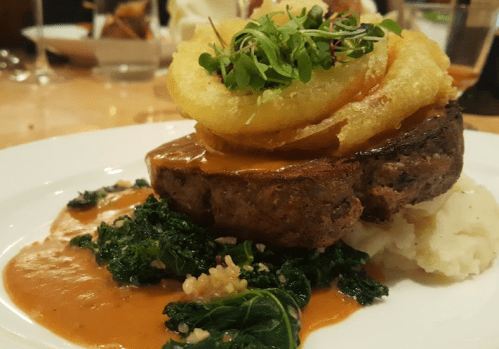 A plated dish featuring meatloaf topped with onion rings, served with mashed potatoes, greens, and a savory sauce.