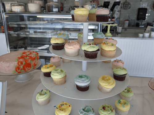 A tiered display of colorful cupcakes in various flavors, with a cake in the background at a bakery.
