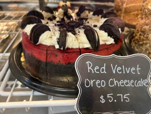 A red velvet Oreo cheesecake topped with cream and cookie pieces, displayed in a bakery case with a price tag.