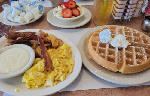A plate with a waffle topped with whipped cream, scrambled eggs, bacon, and grits, alongside a bowl of fruit.