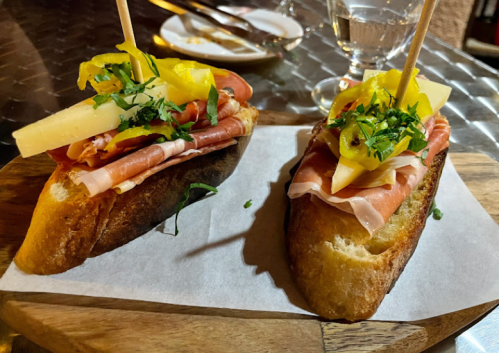 Two slices of toasted bread topped with cured meats, cheese, pickled peppers, and fresh herbs on a wooden board.