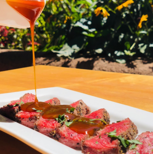 A plate of sliced steak drizzled with sauce, garnished with herbs, set against a backdrop of flowers.