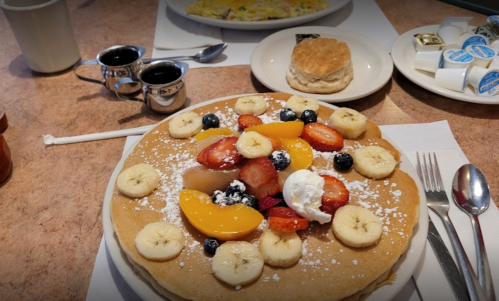 A stack of pancakes topped with bananas, strawberries, blueberries, and peaches, served with syrup and a biscuit.