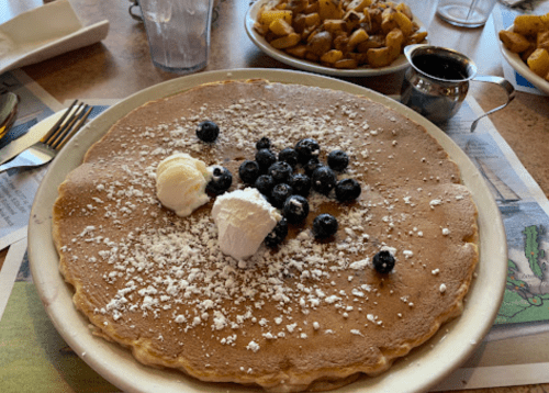 A large pancake topped with blueberries and whipped cream, served with a side of golden-brown potatoes.