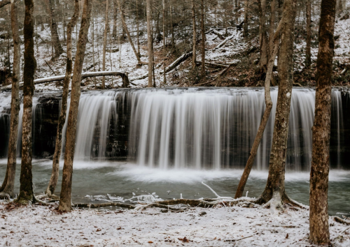 A serene waterfall cascades over rocks, surrounded by trees and a dusting of snow in a tranquil forest setting.