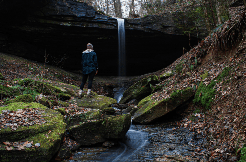 A person stands on mossy rocks by a stream, gazing at a waterfall beneath a rocky overhang in a forested area.