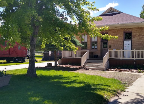 A brick building with a porch, surrounded by green grass and trees, on a sunny day. A red train car is visible in the background.