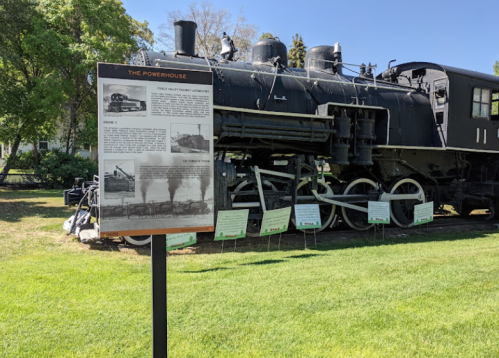 A historic steam locomotive displayed outdoors next to an informational sign about its history. Green grass surrounds the area.