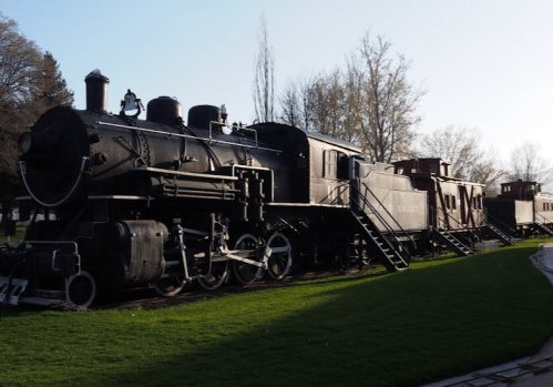 A vintage steam locomotive and train cars displayed on green grass, surrounded by trees in a park setting.