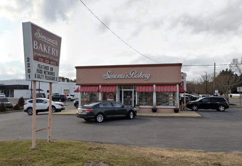 A bakery storefront with a sign, featuring a parking lot and nearby road, under a cloudy sky.