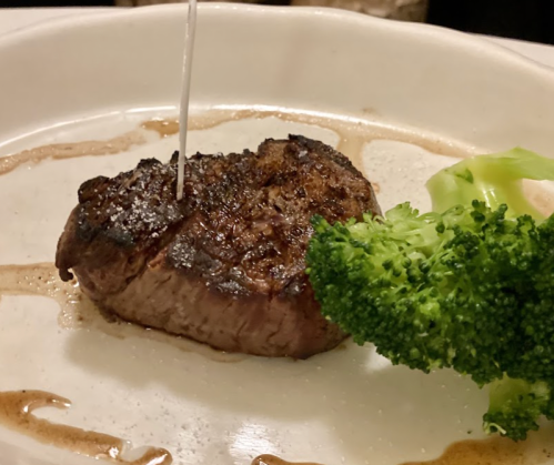 A perfectly cooked steak drizzled with sauce, accompanied by a piece of broccoli on a white plate.