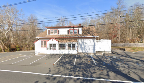 A small, two-story building with a brown roof and white exterior, surrounded by empty parking spaces and trees.