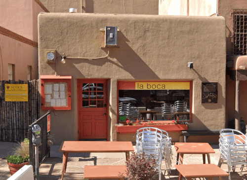 A small adobe-style restaurant with a red door and outdoor seating, featuring a sign that reads "la boca."