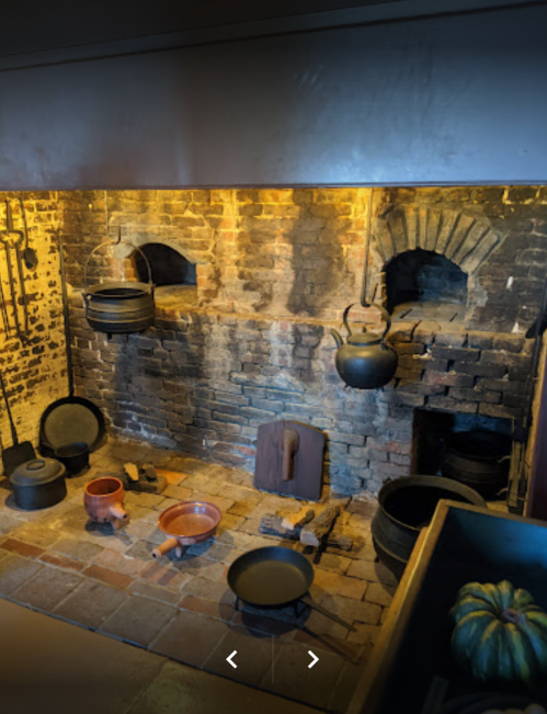 A rustic kitchen with brick walls, hanging pots, and various cooking utensils on a tiled floor.