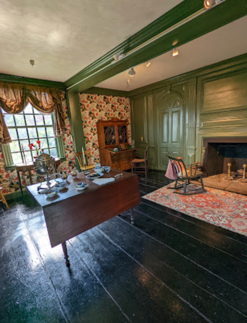 A vintage room with floral wallpaper, a wooden table set with dishes, a fireplace, and period furniture.