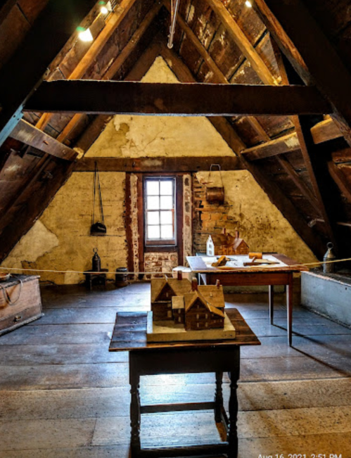 A rustic attic with exposed beams, featuring tables and a model house, illuminated by natural light from a window.
