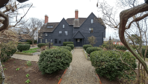 A large black house with a steep roof, surrounded by manicured gardens and gravel pathways on a cloudy day.