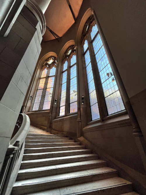 A winding staircase leads up to large stained glass windows, casting colorful light in a historic building.