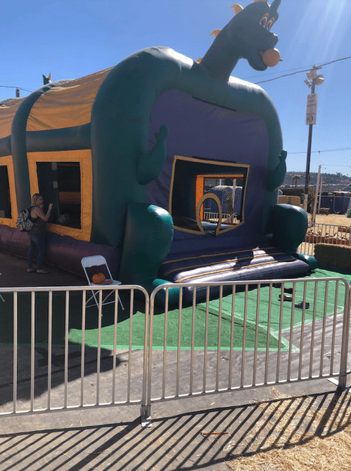 A colorful dragon-themed bounce house with a green and purple design, surrounded by a white fence at an outdoor event.