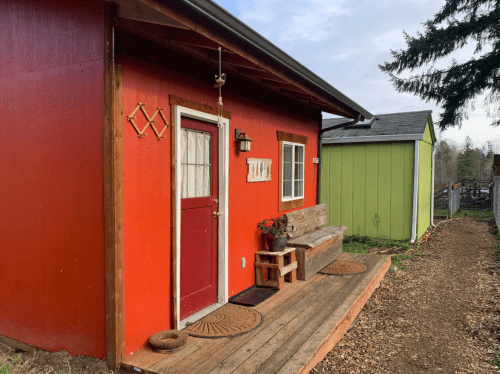 A bright red cabin with a wooden porch, potted plants, and a green building in the background.