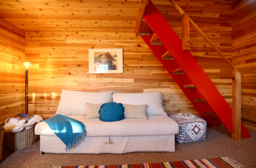 Cozy wooden interior with a sofa, colorful rug, and a red staircase leading to an upper level. Warm lighting enhances the space.