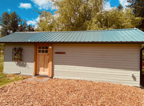 A small, light-colored cabin with a green metal roof, surrounded by trees and wood chips on the ground.