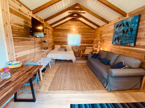 Cozy wooden interior of a cabin with a bed, sofa, dining table, and decorative artwork on the walls.