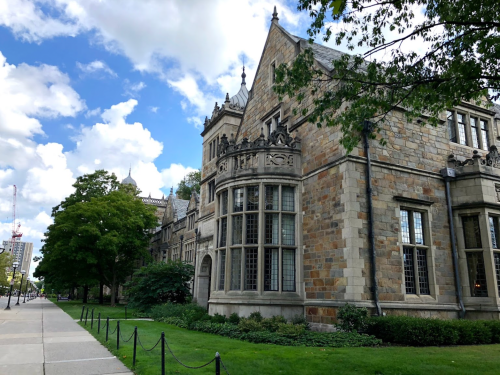 Historic stone building with large windows, surrounded by greenery and a sidewalk under a partly cloudy sky.