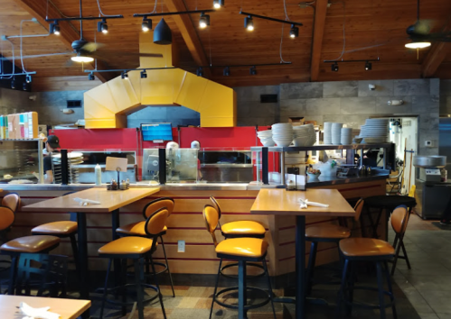 Interior of a restaurant featuring wooden beams, a large oven, and empty tables with chairs.