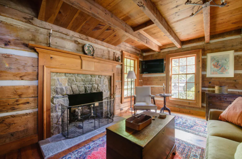 Cozy rustic living room with wooden beams, stone fireplace, and warm lighting, featuring a comfortable chair and colorful rug.