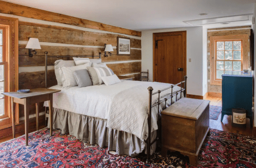 Cozy bedroom with a wooden bed, rustic walls, and a colorful rug, featuring natural light from windows.