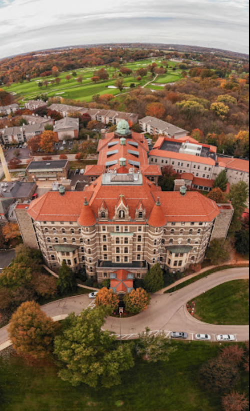 Aerial view of a large historic building with red roofs, surrounded by trees and a golf course in autumn colors.
