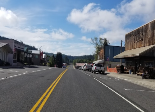 A quiet street in a small town, lined with buildings and parked cars under a blue sky with scattered clouds.