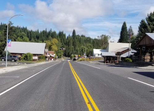 A quiet road lined with small buildings and trees, leading into a forested area under a partly cloudy sky.