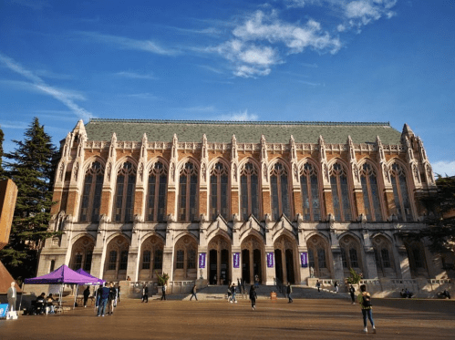 A large, historic brick building with Gothic architecture, set against a blue sky, with people walking in front.