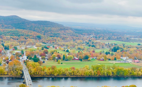 Aerial view of a colorful autumn landscape with a river, bridge, and small town surrounded by hills.