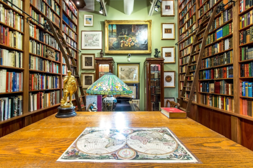 A cozy library filled with books, featuring a wooden table, a globe map, and decorative lamps amidst tall shelves.