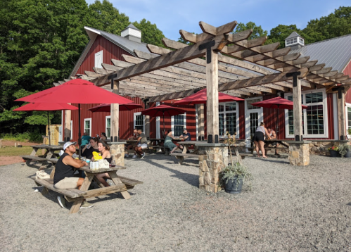 Outdoor dining area with wooden tables, red umbrellas, and a rustic red building in the background surrounded by greenery.