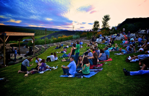 A crowd enjoys an outdoor event on a grassy hill at sunset, with chairs and blankets scattered around.