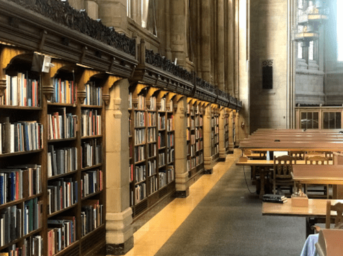 A spacious library interior with tall bookshelves, wooden tables, and large windows allowing natural light.