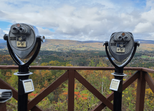 Two binoculars on a viewing platform overlooking a colorful autumn landscape with mountains and clouds in the background.