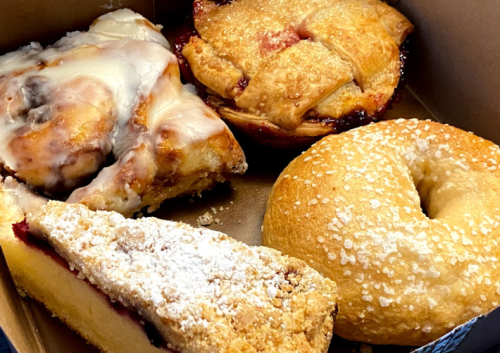 A box containing a cinnamon roll, fruit pie, crumb cake, and a plain bagel, all freshly baked and appetizing.