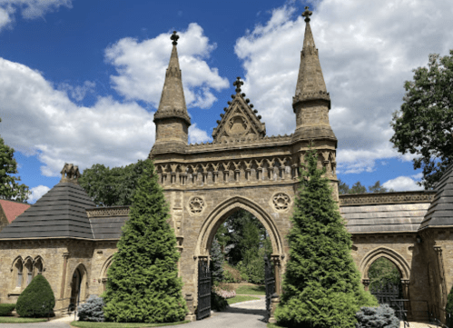 A grand stone gate with tall spires, flanked by lush greenery, under a blue sky with fluffy clouds.